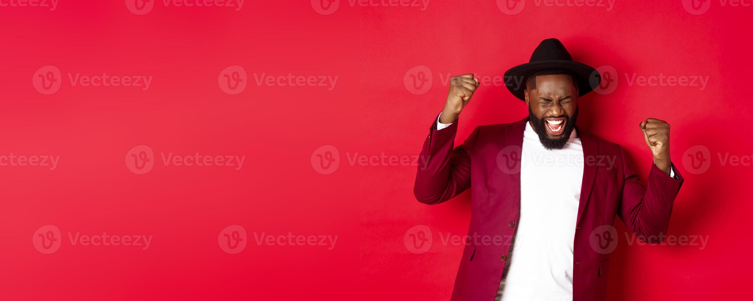 Successful Black man in suit saying yes, rejoicing of winning or achieving goal, raising hands up and screaming of joy, red background photo
