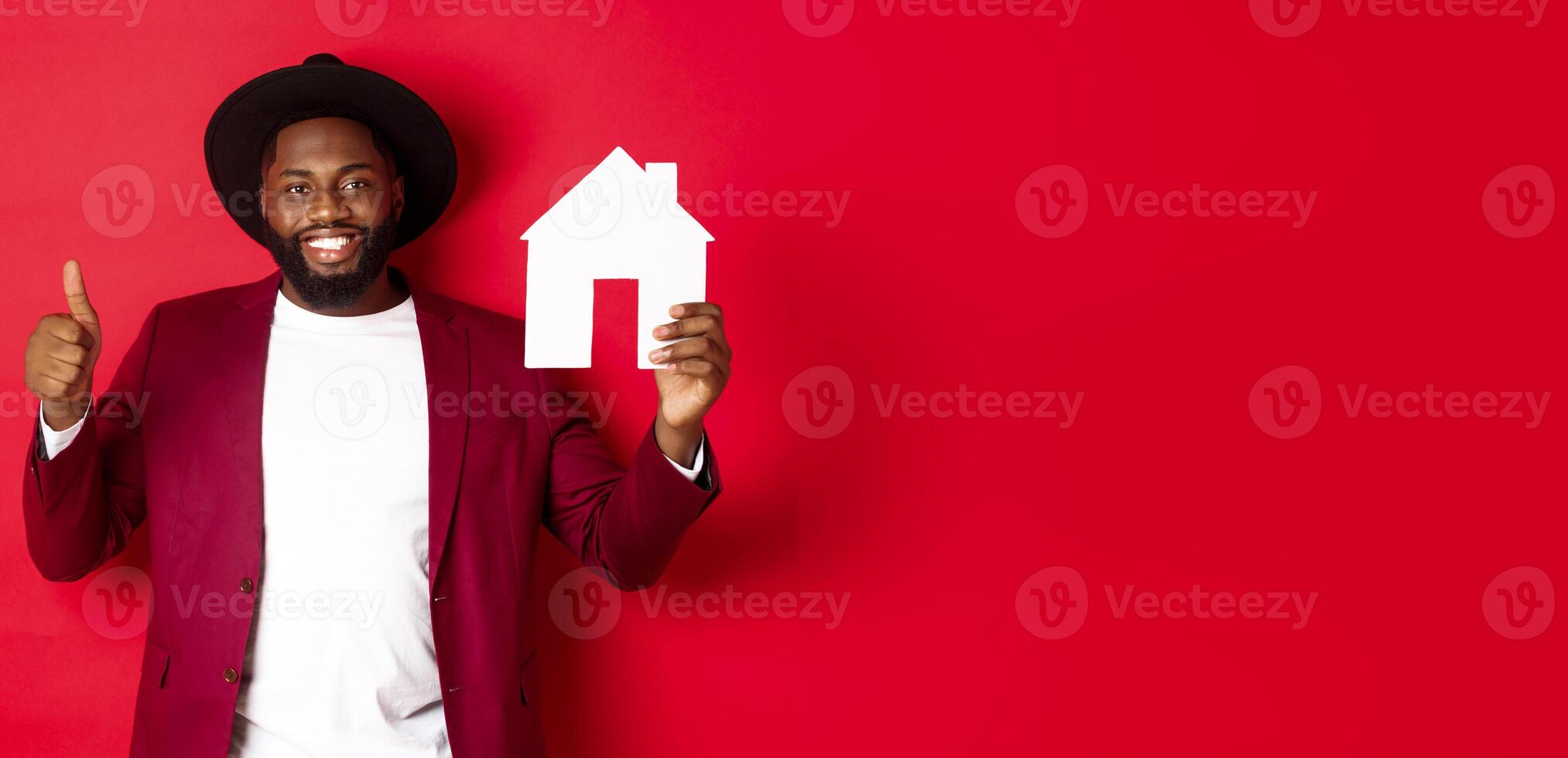 Real estate. Stylish african american bearded man showing home maket, thumb up, recommending agency or apartment complex, standing over red background photo