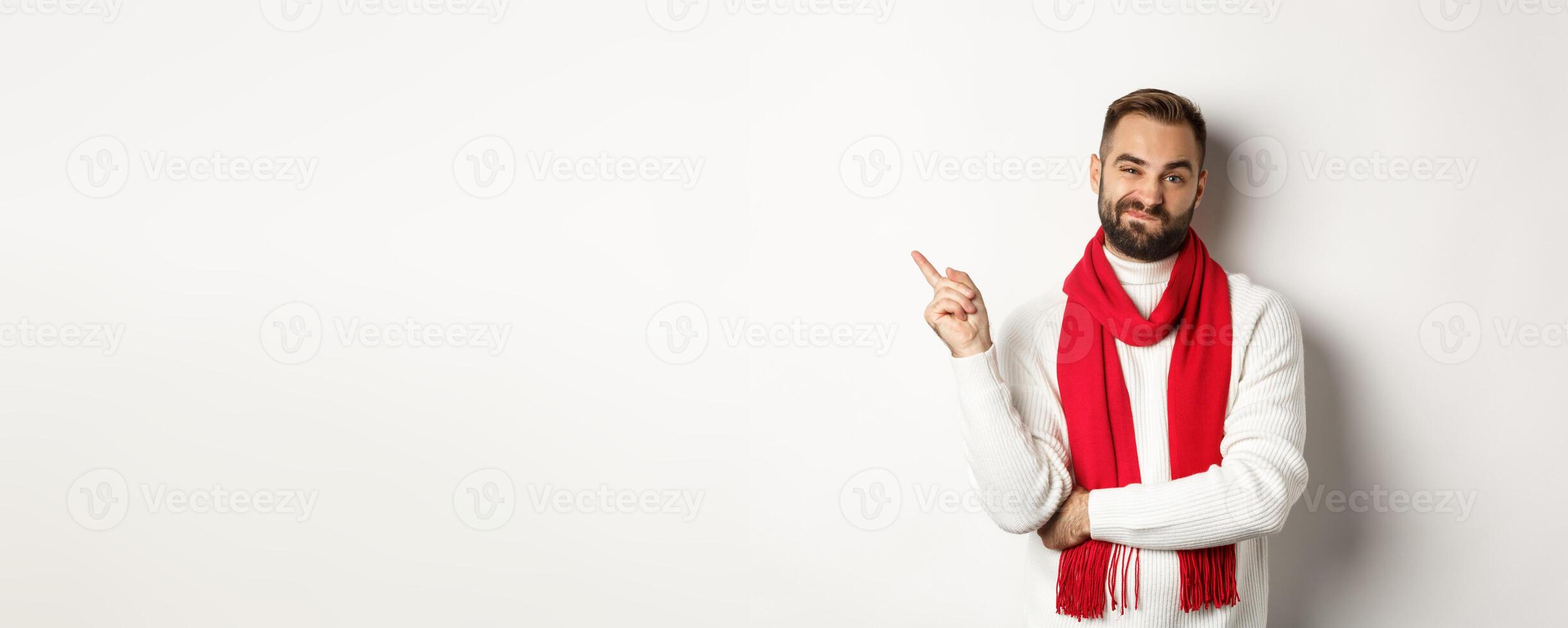 Christmas shopping and winter holidays concept. Skeptical young man showing something bad, pointing finger left and grimacing displeased, standing over white background photo