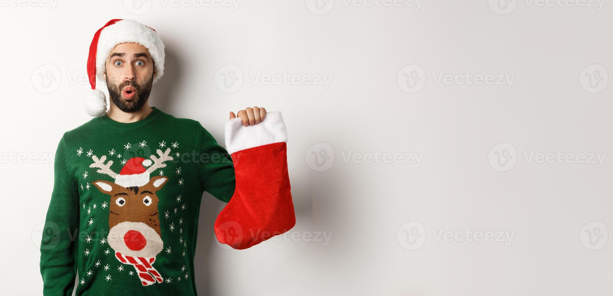 Xmas and winter holidays concept. Happy man got a gift in Christmas sock, looking excited, standing in Santa hat against white background photo