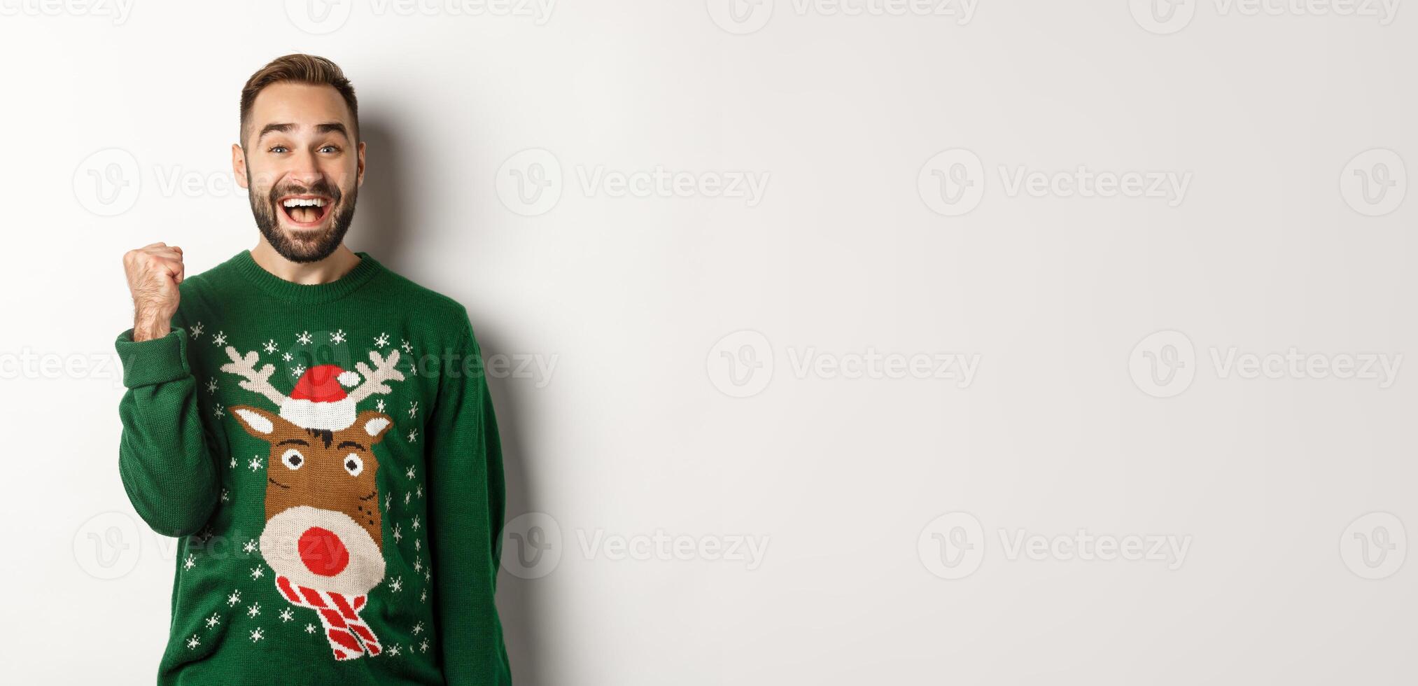 New Year, holidays and celebration. Excited and happy guy in Christmas sweater making fist pump, triumphing, receiving a gift and rejoicing, standing over white background photo