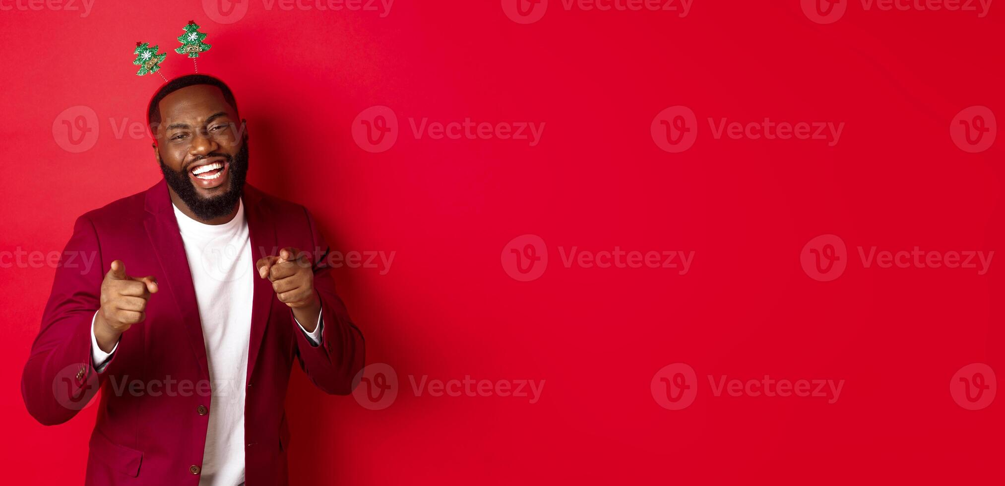 Merry Christmas. Handsome african american man in blazer and party headband, celebrating new year, pointing at camera and smiling, congratulating, red background photo