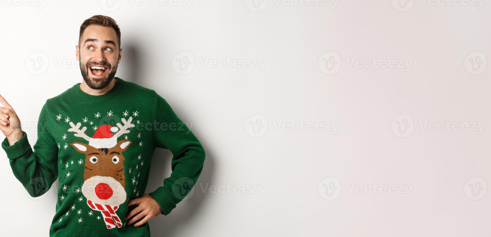 Winter holidays and christmas. Happy man in funny sweater pointing finger at upper left corner logo, showing advertisement, white background photo