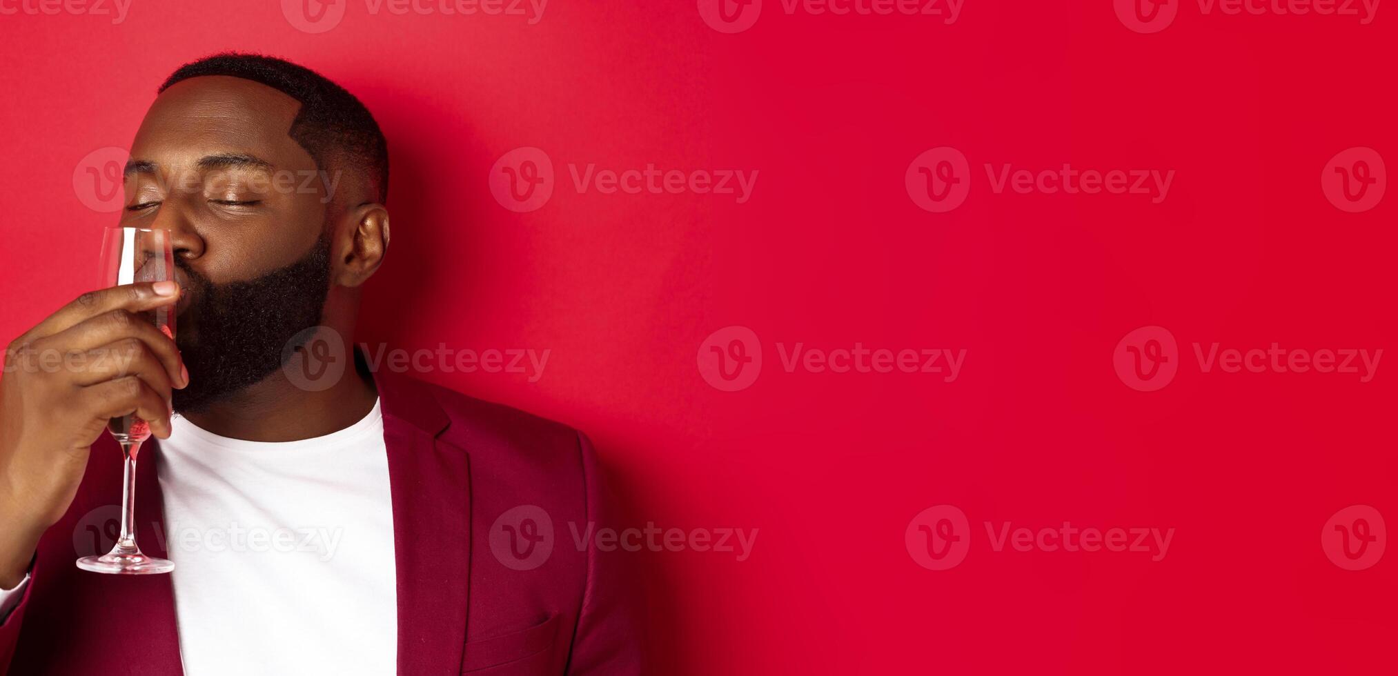 Drunk african american guy kissing glass with champagne, celebrating new year, enjoying party, standing over red background photo