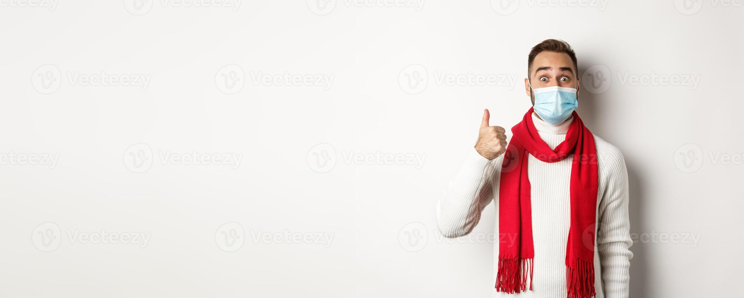 Covid-19, lockdown and quarantine concept. Amazed and satisfied man buyer in medical mask showing thumb-up, looking excited, like something good, white background photo