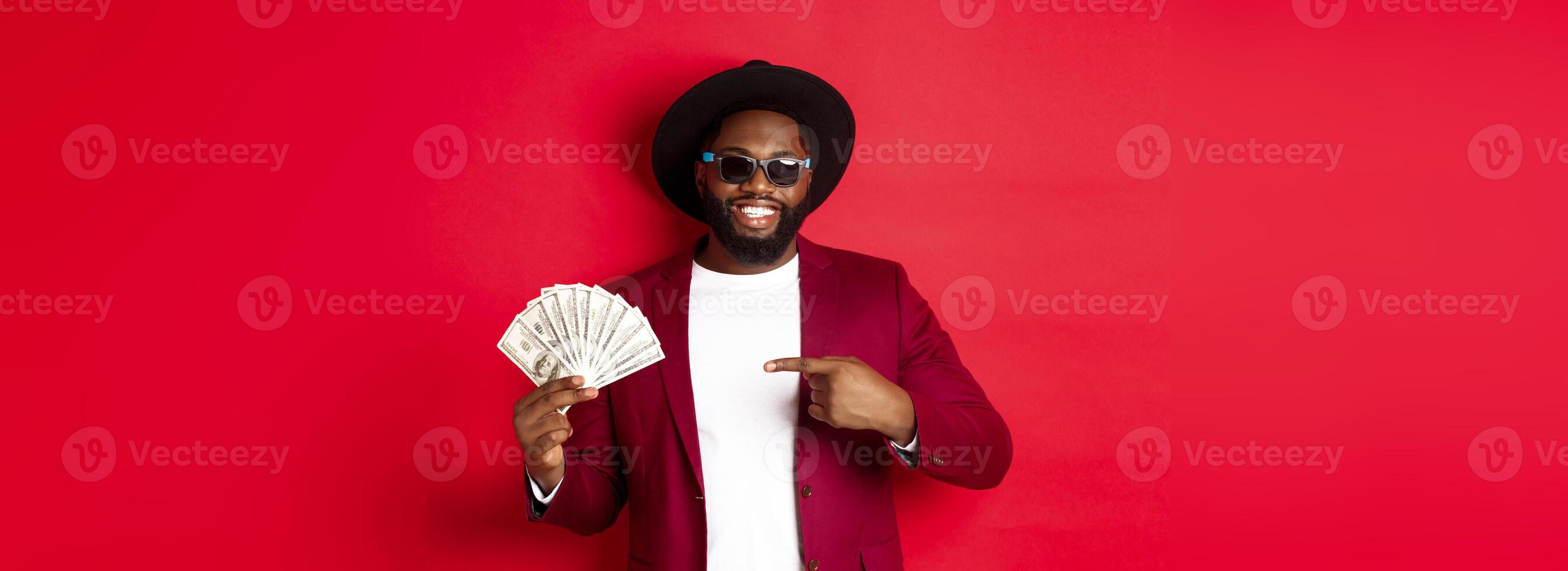 Handsome and stylish african american male model showing money and smiling, wearing sunglasses and fancy hat, standing over red background photo