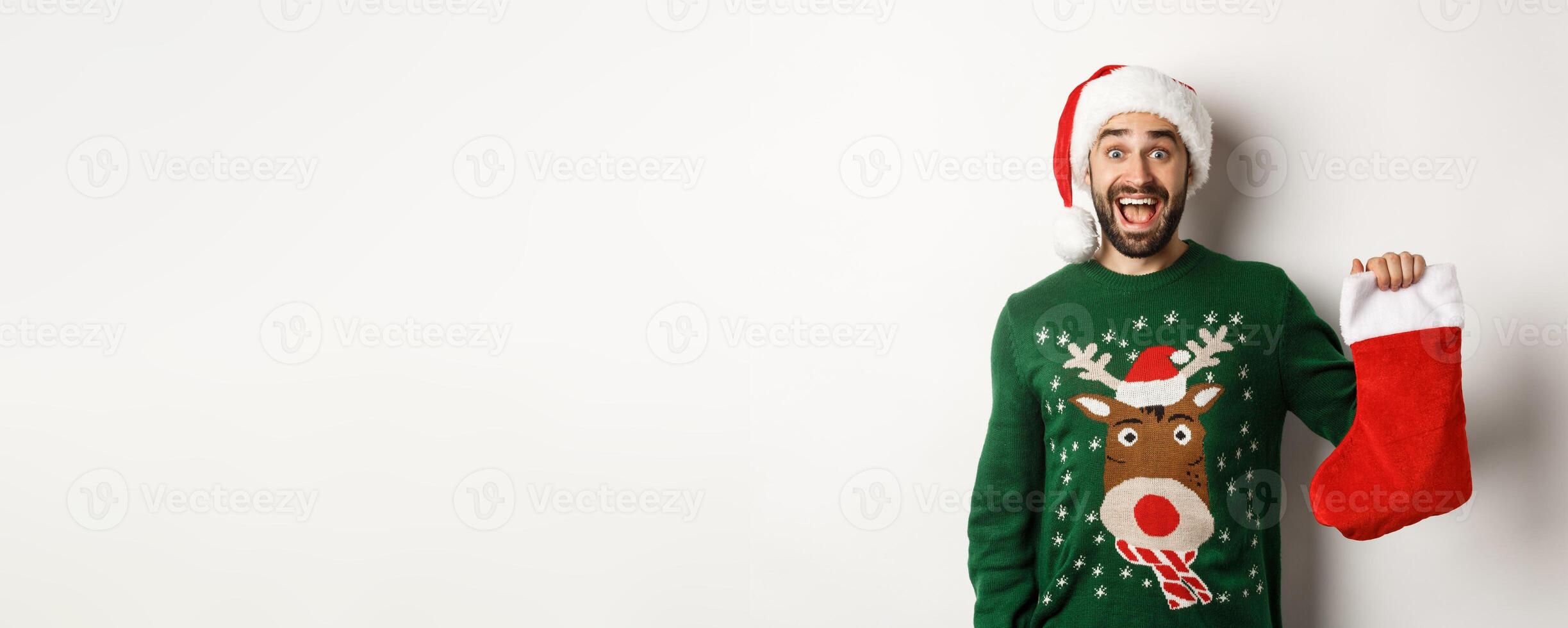 Xmas and winter holidays concept. Happy man got a gift in Christmas sock, looking excited, standing in Santa hat against white background photo