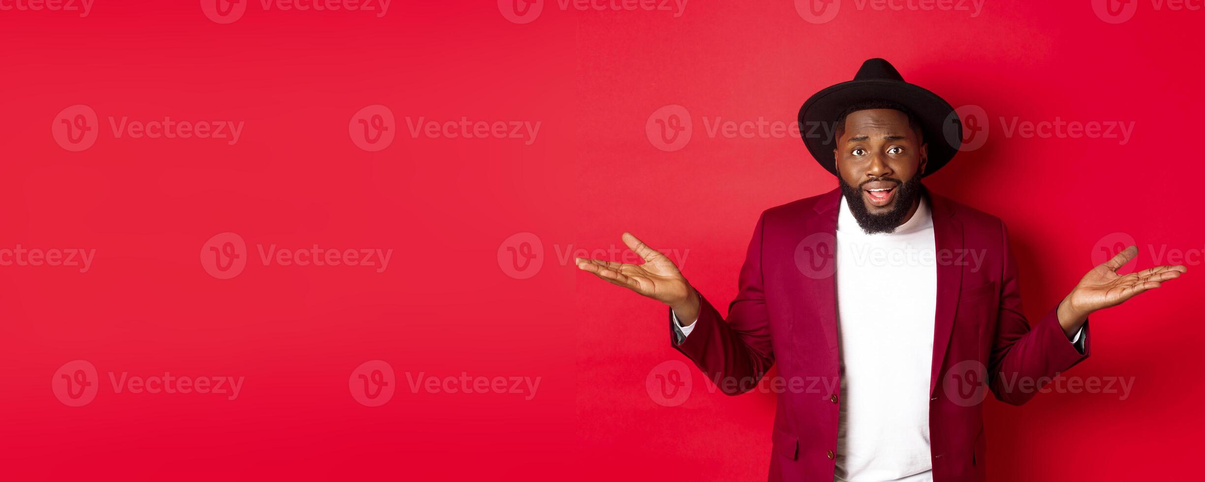 Image of confused Black man asking question, spread hands sideways and staring camera clueless, standing against red background photo