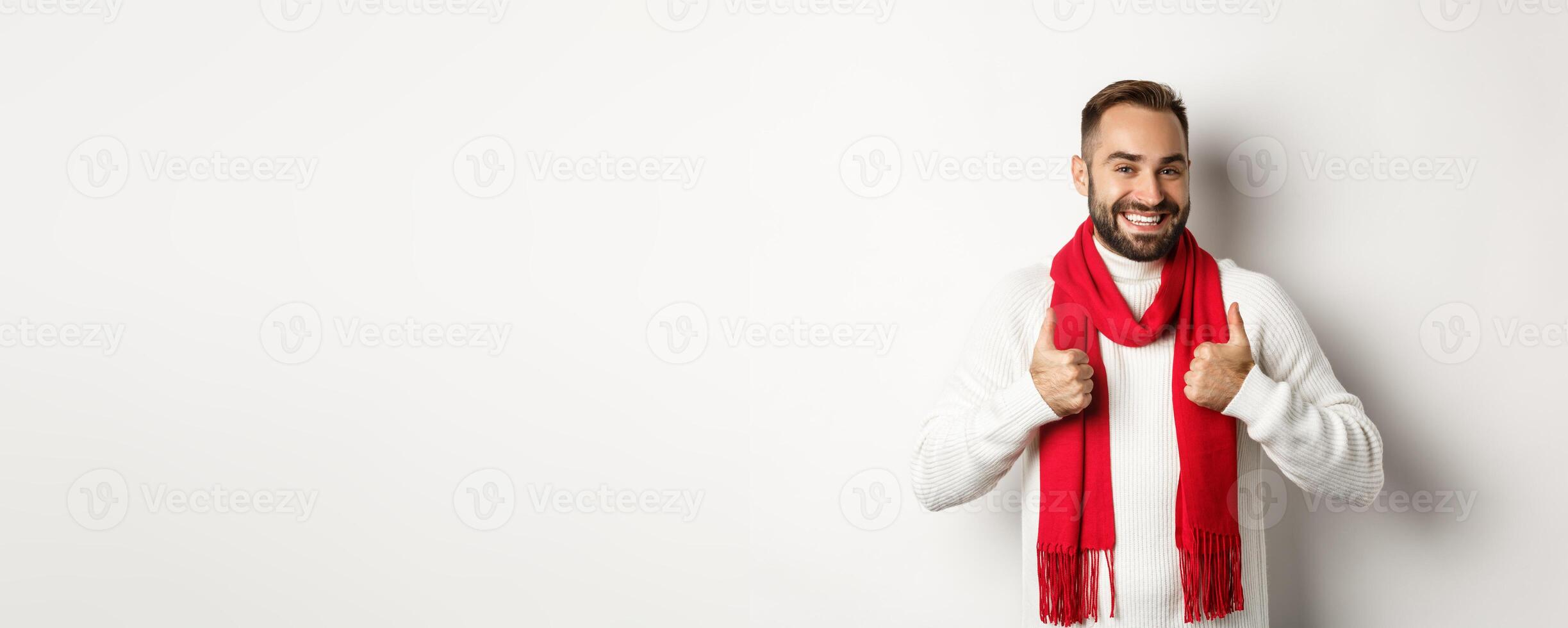Winter holidays and shopping concept. Cheerful bearded man in red christmas scarf showing thumbs up, approve and recommend store, standing over white background photo