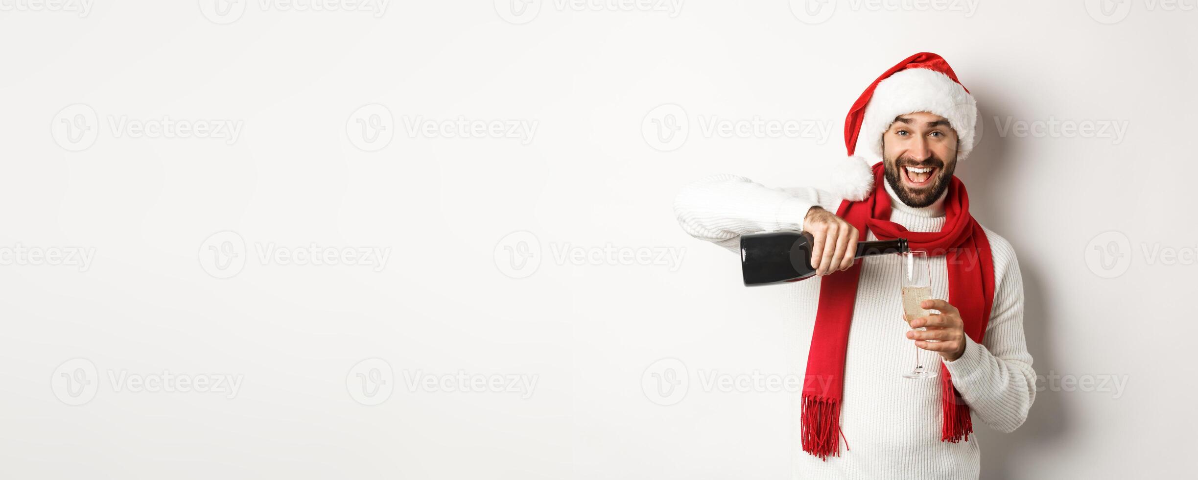 Christmas party and holidays concept. Happy bearded man in Santa hat and scard, pour himself champagne and smiling, celebrating New Year, standing over white background photo
