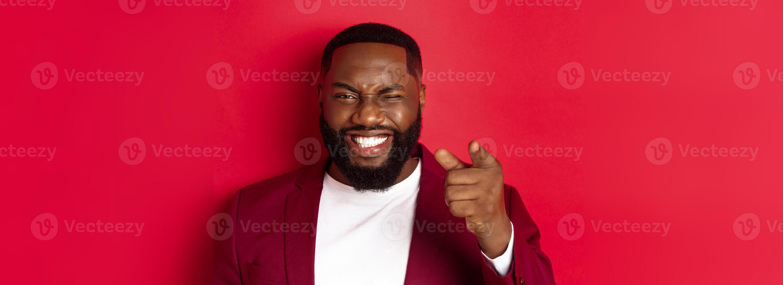 Cheerful Black man with beard wearing party clothes, pointing finger at camera and winking, congratulate with christmas and new year, standing over red background photo