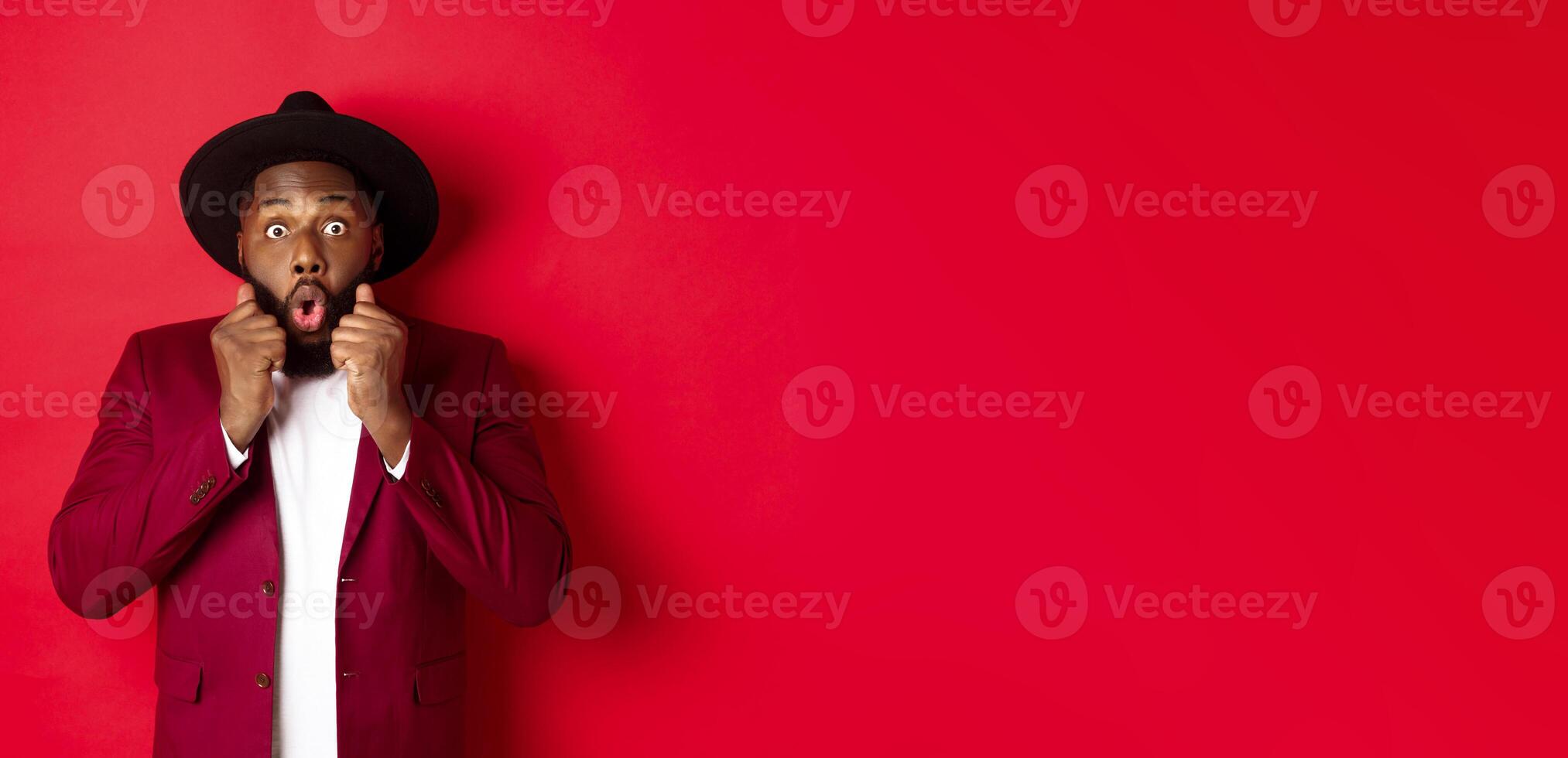 Fashion and party concept. Surprised african american male model looking shocked and amazed, saying wow and staring camera, standing against red background photo