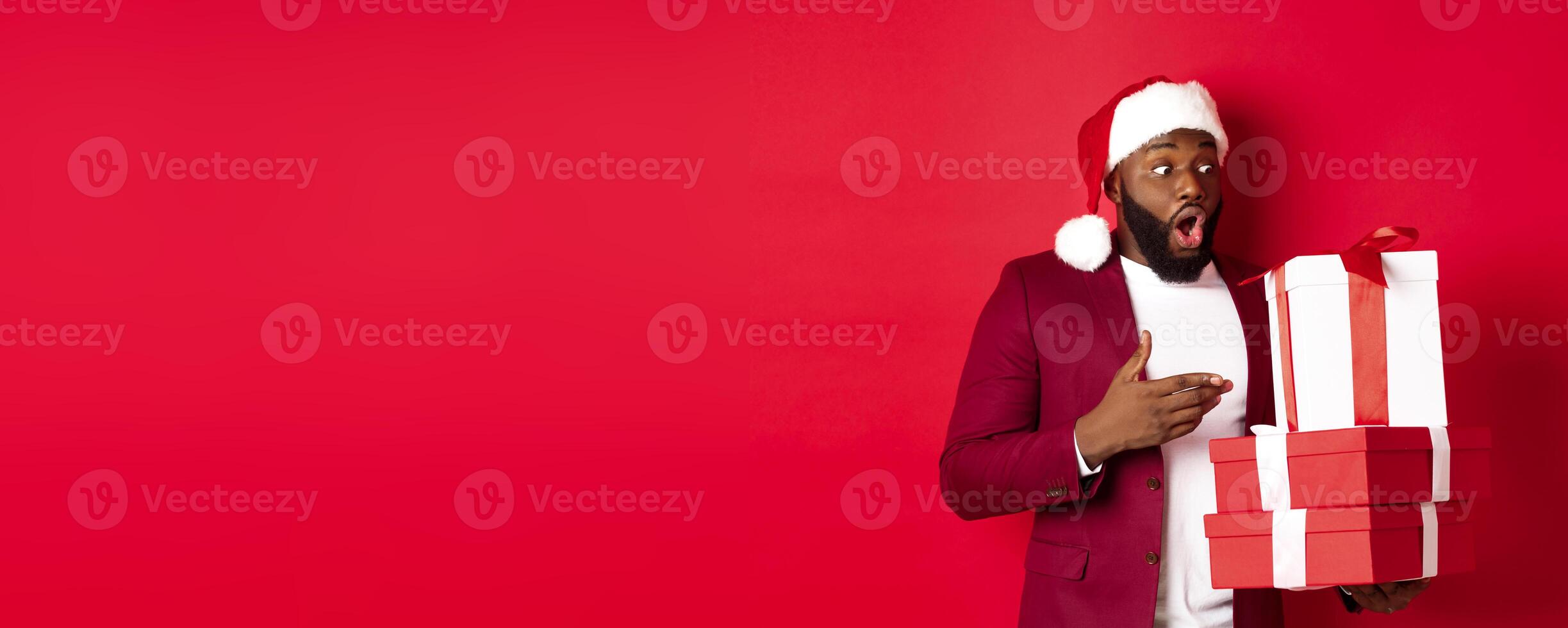 Christmas, New Year and shopping concept. Surprised Black man looking at xmas presents with amazement, wearing santa hat, standing with gifts against red background photo