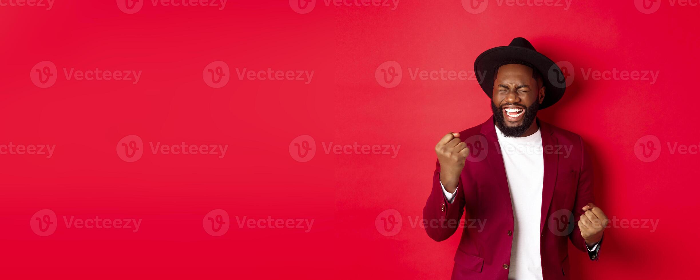 Lucky african american man celebrating victory, winning and rejoicing with fist pumps, screaming happy with closed eyes, red background photo