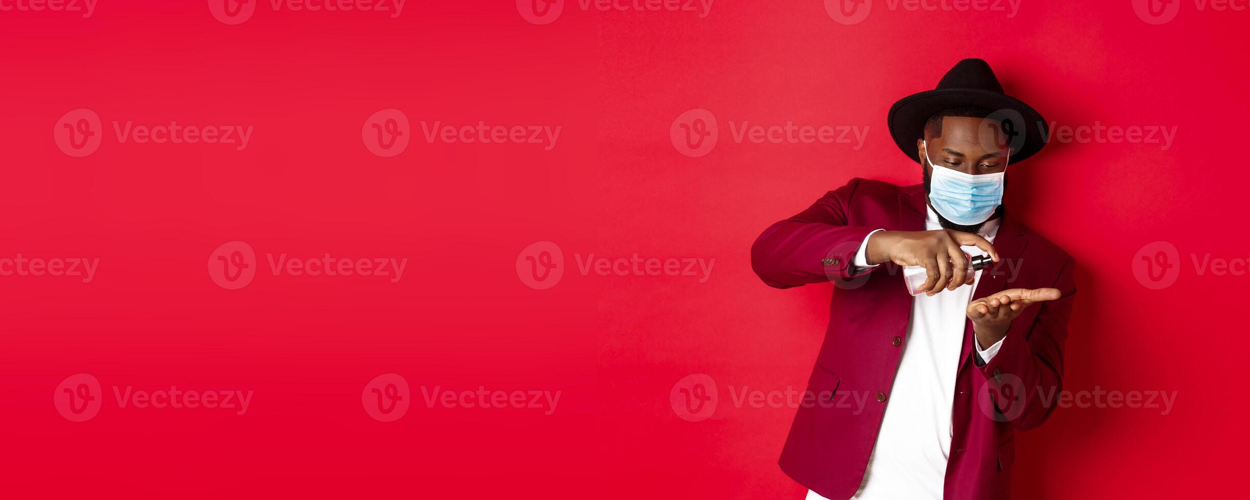 Covid-19, quarantine and holidays concept. Handsome Black man in face mask and party outfit, disinfecting hands with hand sanitizer, using antiseptic, standing over red background photo