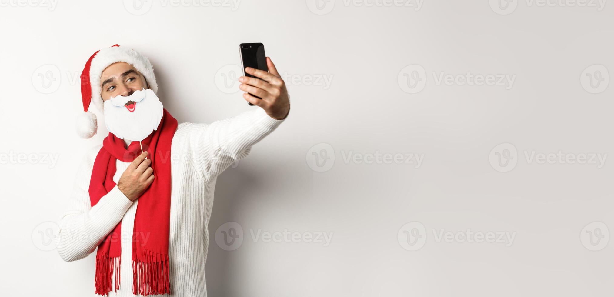 Christmas party and celebration concept. Young man taking selfie with funny white beard Santa mask and hat, posing for photo on mobile phone, studio background