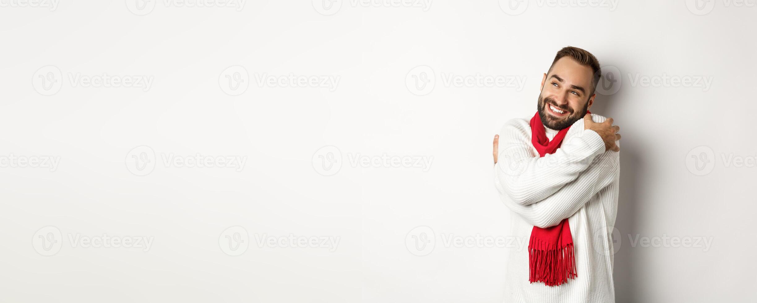 Happy smiling man hugging himself and looking satisfied at upper left corner, wearing warm and comfortable winter sweater, standing over white background photo