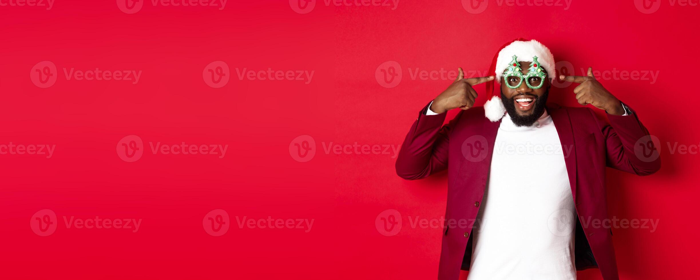 Merry Christmas. Cheerful Black man wearing funny party glasses and santa hat, smiling joyful, celebrating winter holidays, standing over red background photo