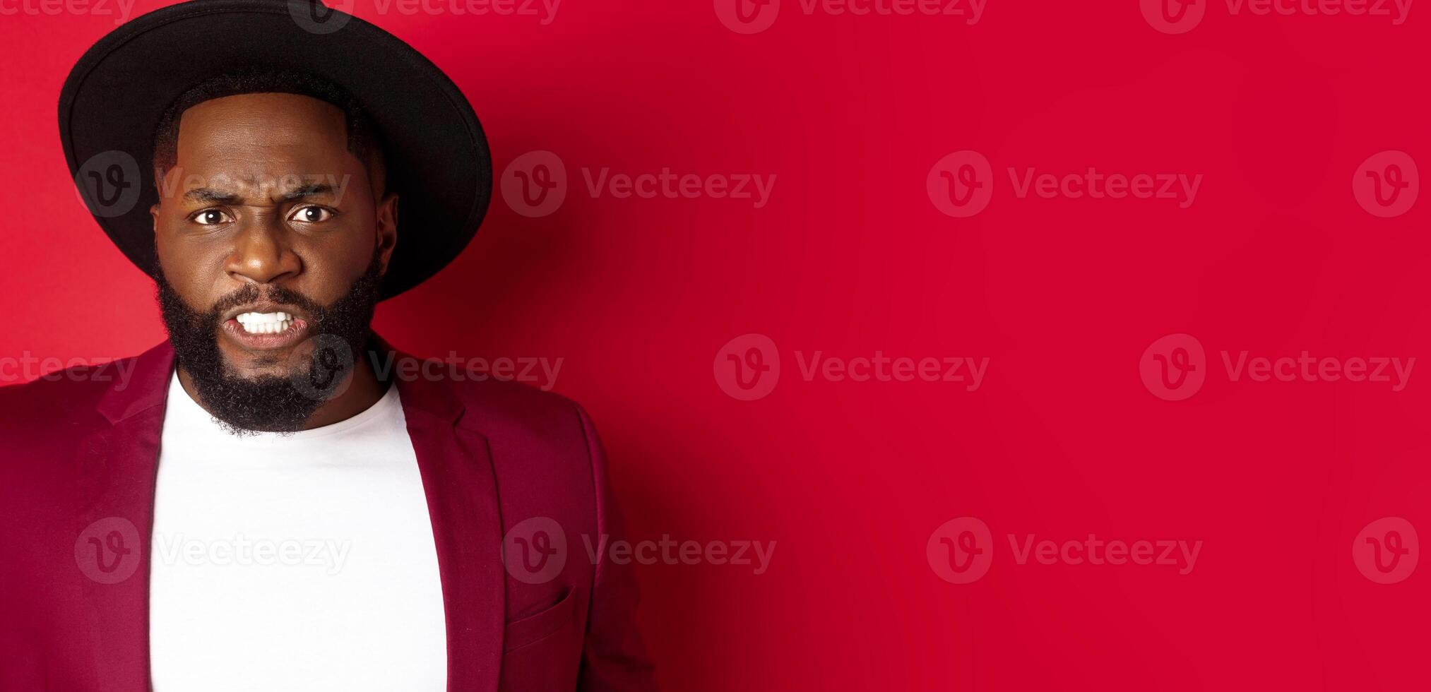 Tensed african american man clench teeth and frowning, looking at something uncomfortable and cringe, standing over red background photo
