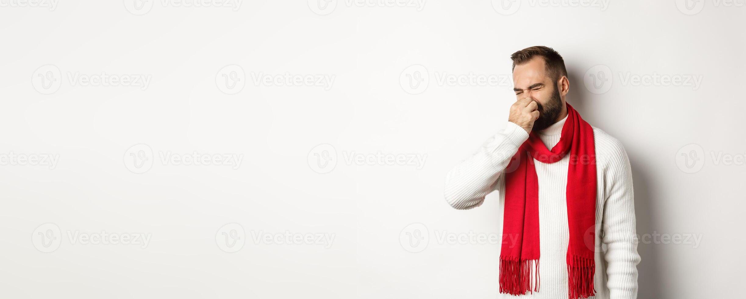 Displeased guy shut his nose from bad smell, grimacing disgusted, standing over white background in white sweater with red scarf photo