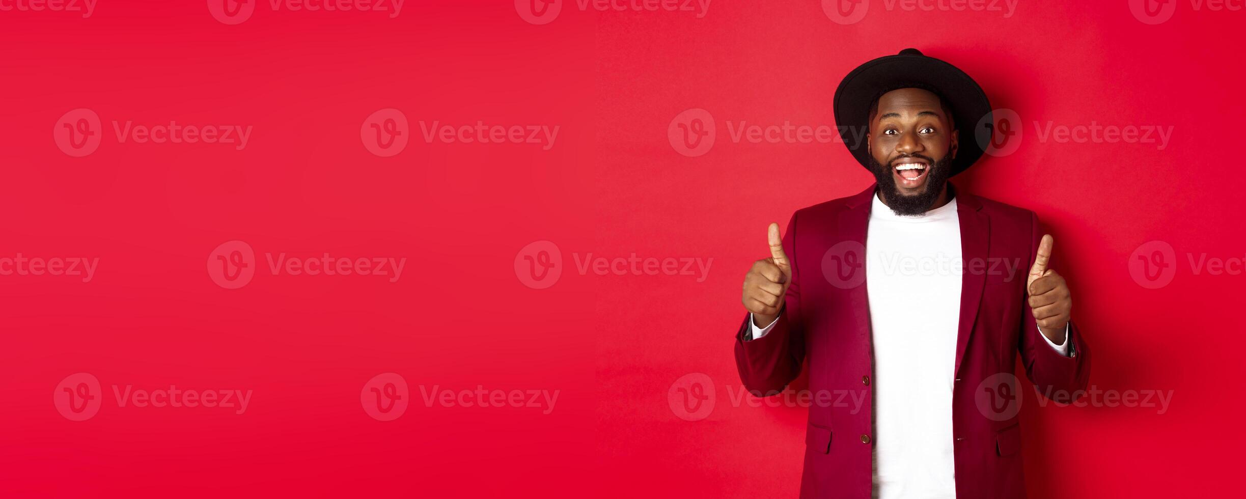 Christmas shopping and people concept. Handsome Black man smiling satisfied, showing thumbs-up, like and agree, approve something, standing against red background photo