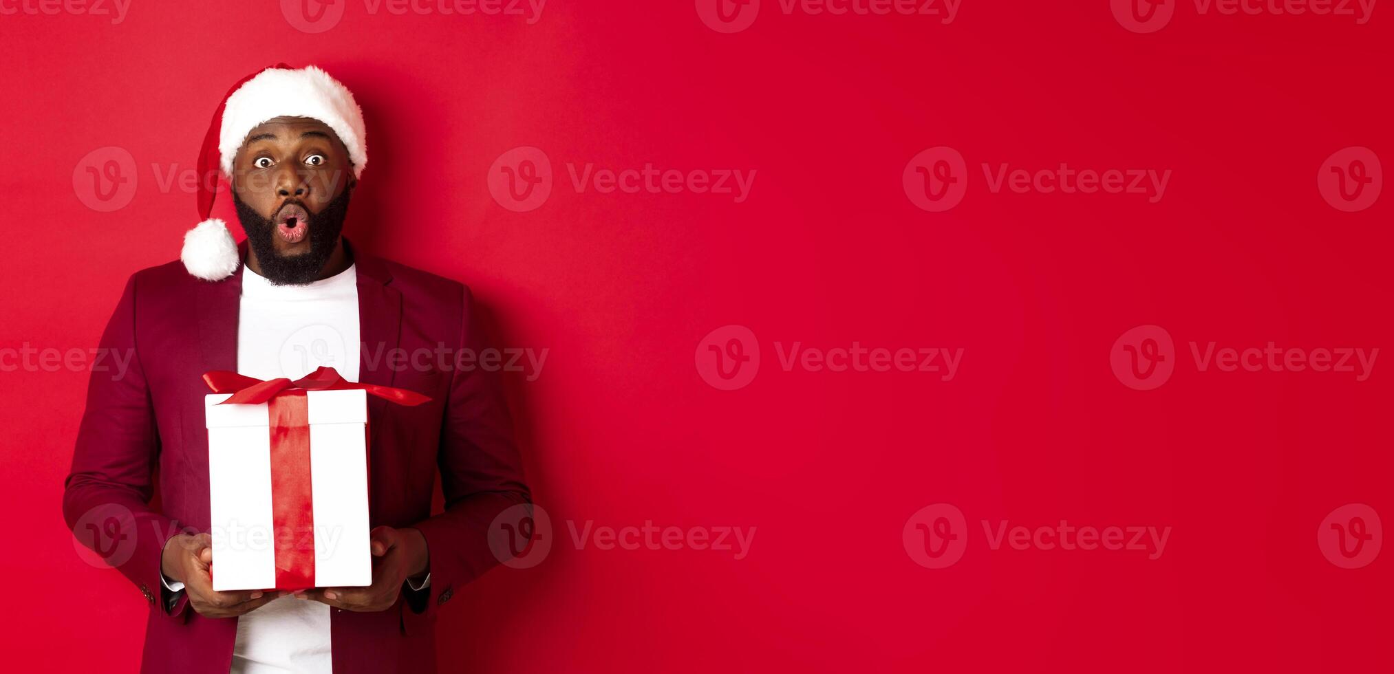 Christmas, New Year and shopping concept. Surprised african american man in santa hat saying wow, holding holiday gift and looking at camera amazed, red background photo