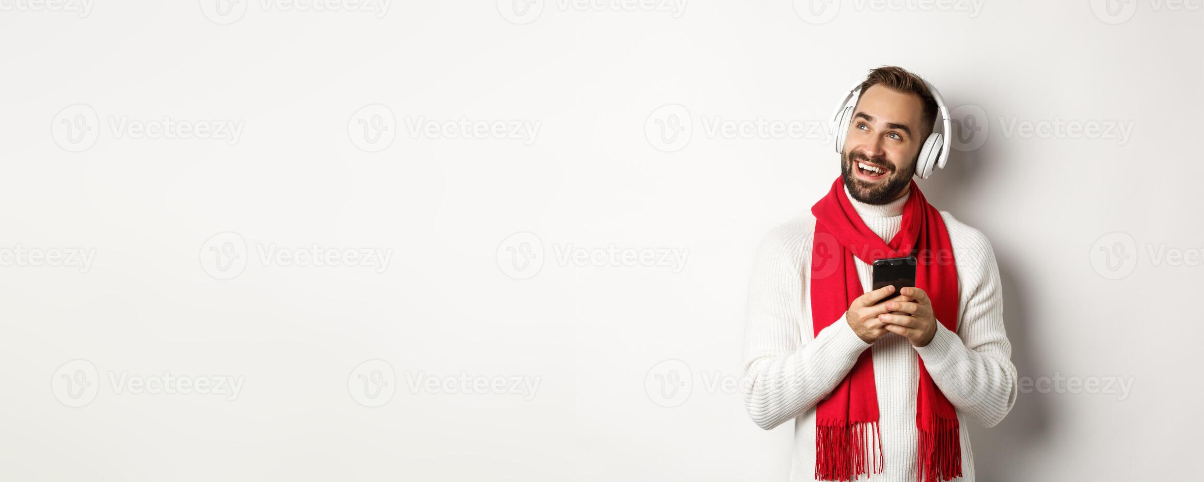 Winter holidays and technology concept. Happy man listening music podcast on headphones, holding mobile phone and looking at empty space, white background photo