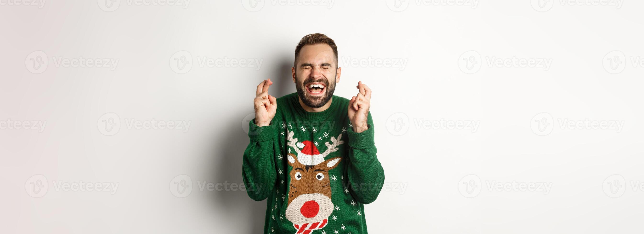 Christmas, holidays and celebration. Hopeful and excited guy making wish, keep fingers crossed for good luck and smiling, standing over white background photo