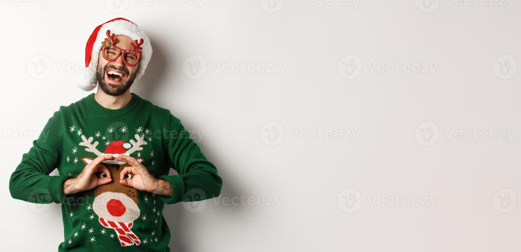 Christmas holidays, celebration concept. Happy guy in Santa hat and party glasses making fun of funny sweater, standing over white background photo