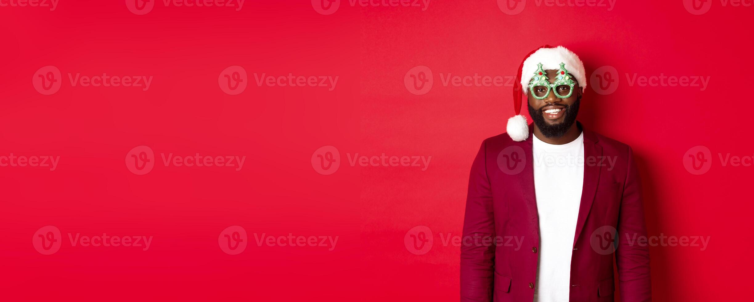 Funny Black man in santa hat and party glasses celebrating Christmas, smiling happy and wishing merry xmas, standing over red background photo