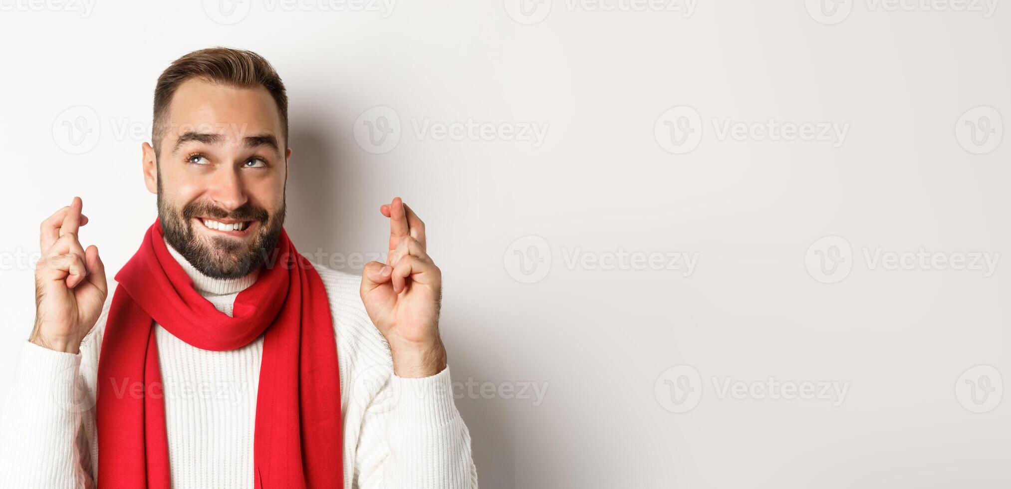 Christmas holidays. Happy attractive man making wish on winter holidays, cross fingers for good luck, looking up, standing over white background photo