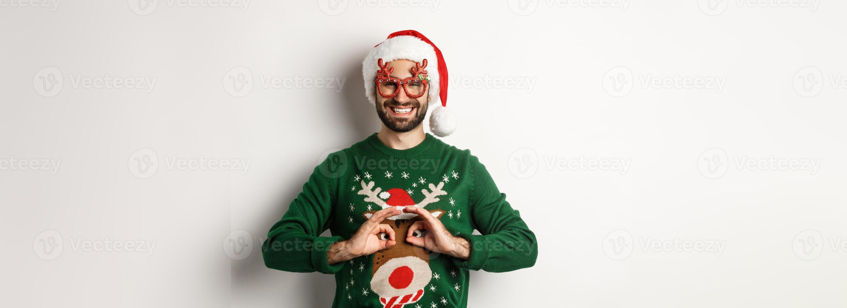Christmas holidays, celebration concept. Happy guy in Santa hat and party glasses making fun of funny sweater, standing over white background photo