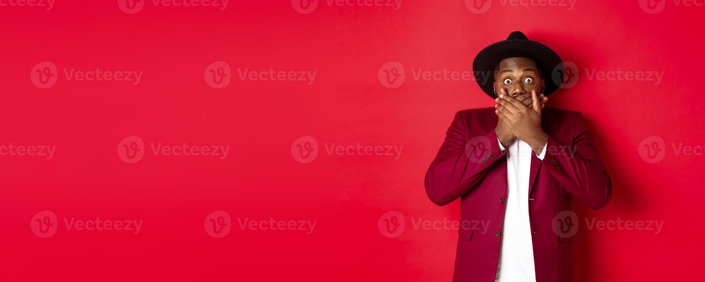 Christmas shopping and people concept. Shocked black man looking startled at camera, covering mouth with hands, standing against red background photo