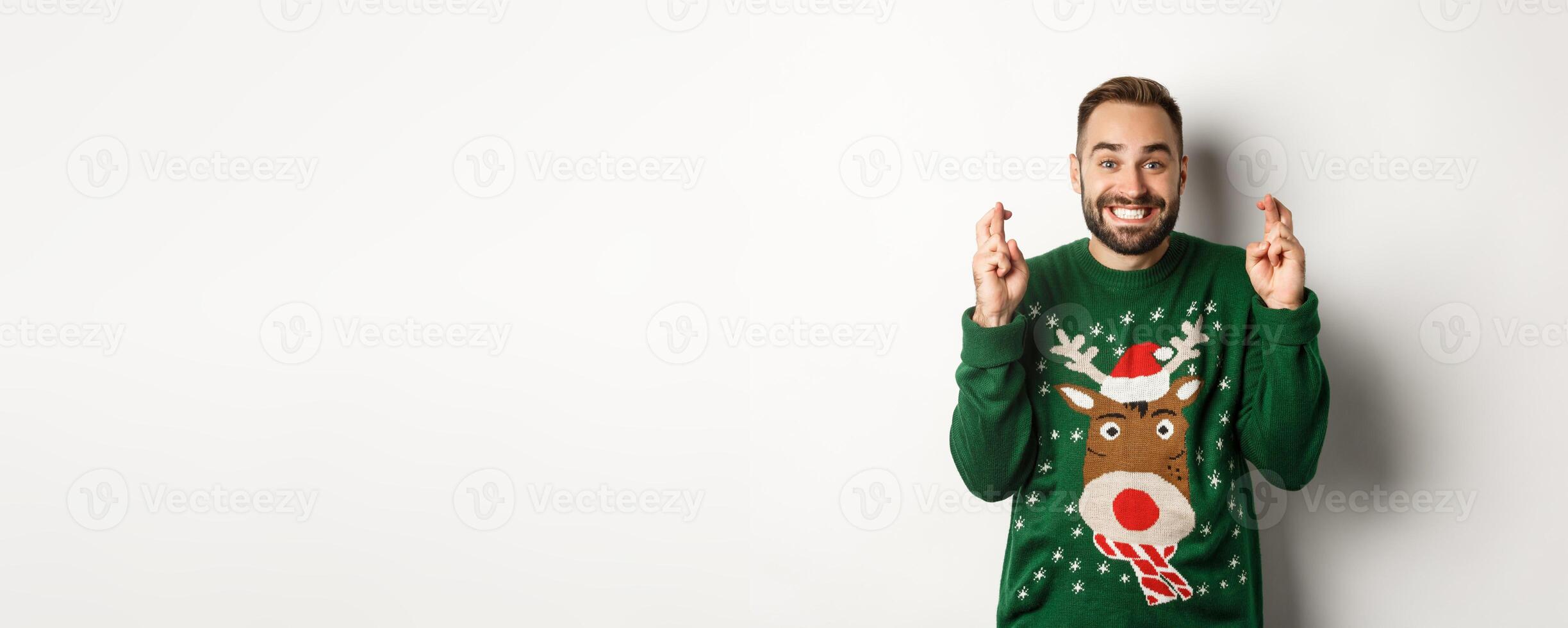 Christmas, holidays and celebration. Hopeful happy guy making wish, keep fingers crossed for good luck and smiling, standing over white background photo