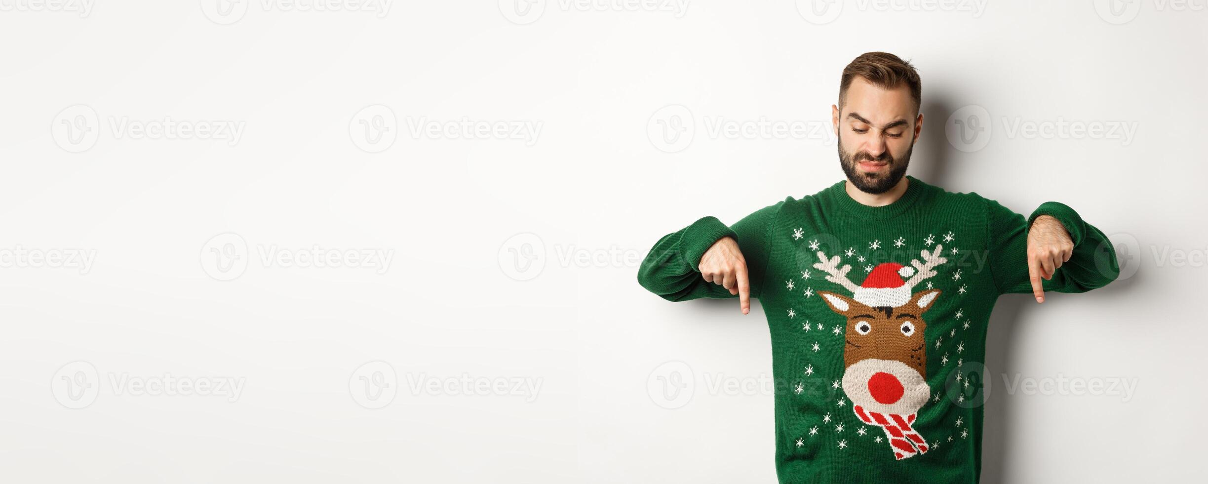 Winter holidays and christmas. Skeptical bearded man in green sweater, pointing and looking down displeased, standing over white background photo
