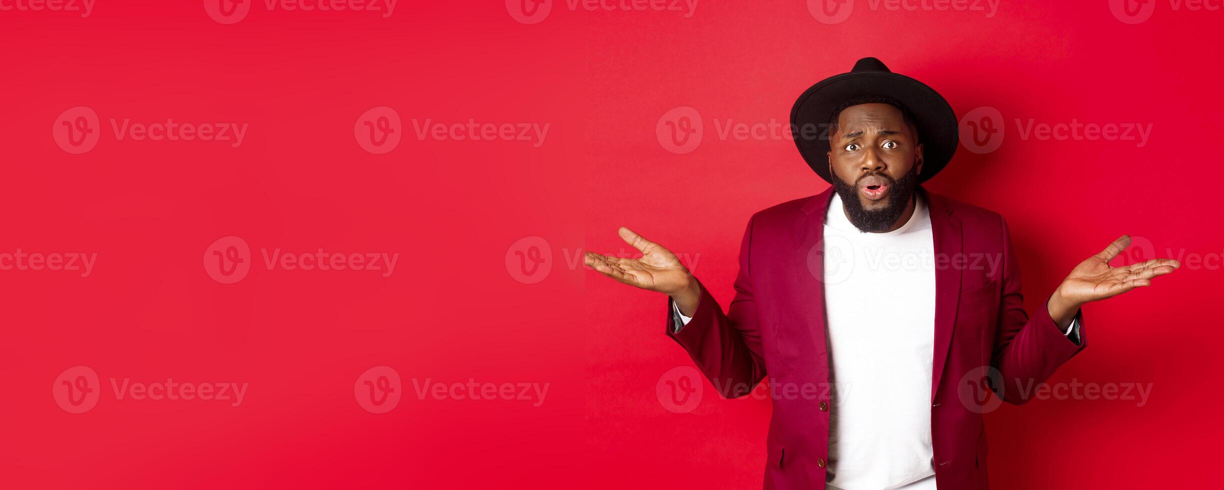 Confused and worried Black man spread hands sideways, staring at camera puzzled, asking what wrong, standing against red background photo
