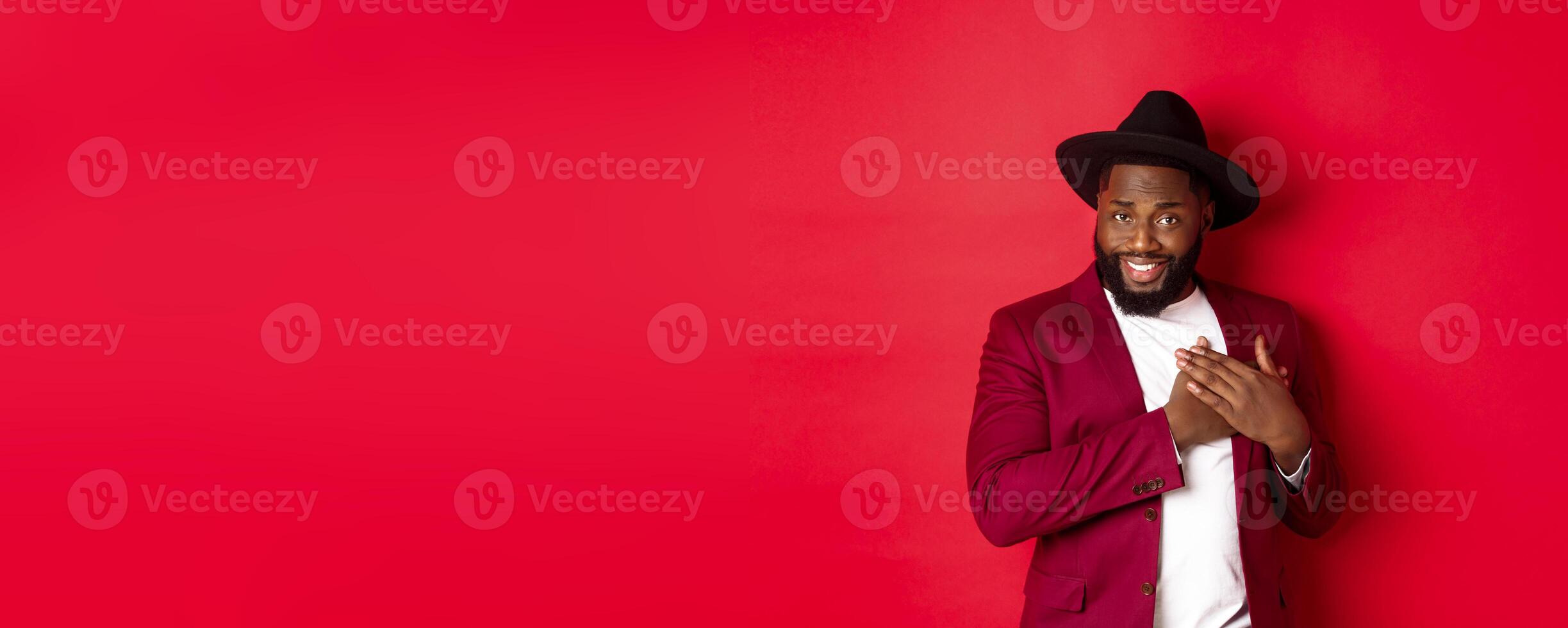 Fashion and party concept. Handsome african american macho man flirting, smiling and holding hands on heart, being grateful and touched, standing over red background photo