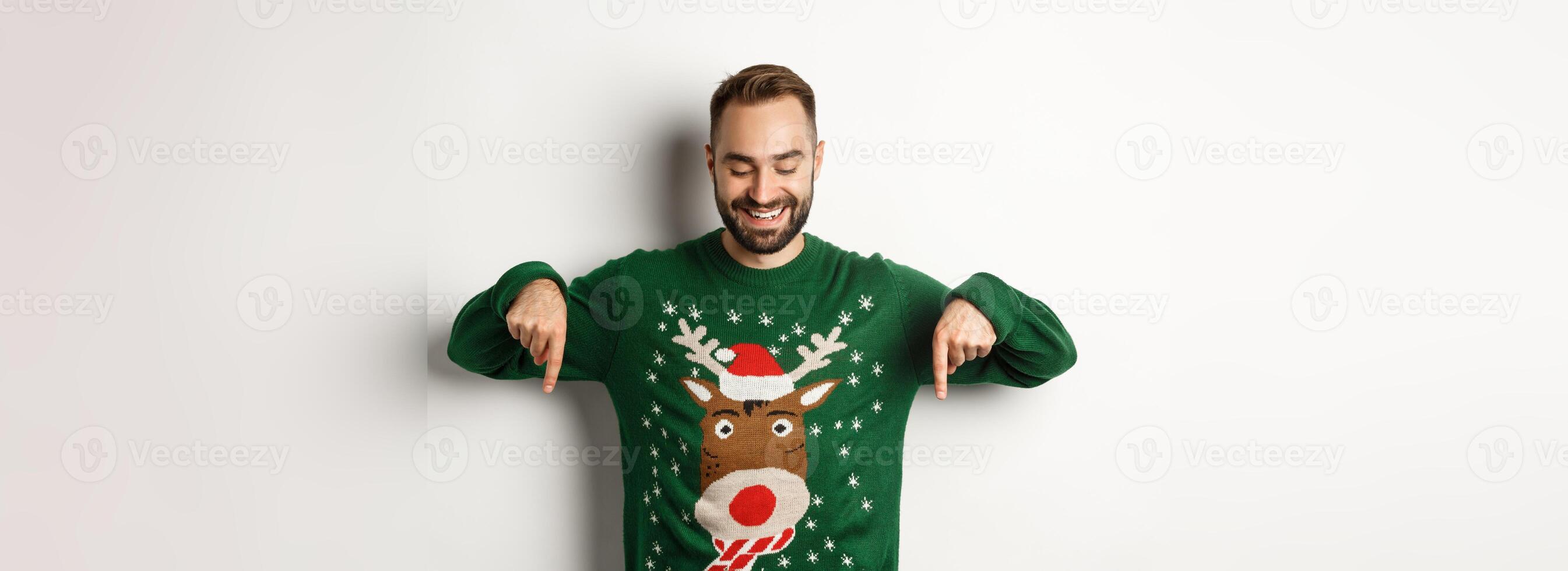 Winter holidays and christmas. Happy caucasian man in green sweater, pointing fingers down and looking satisfied, standing over white background photo