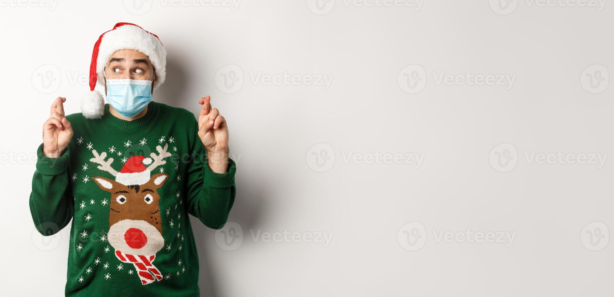 Concept of covid-19 and Christmas holidays. Excited man in face mask and Santa hat cross fingers, making a wish, standing over white background photo