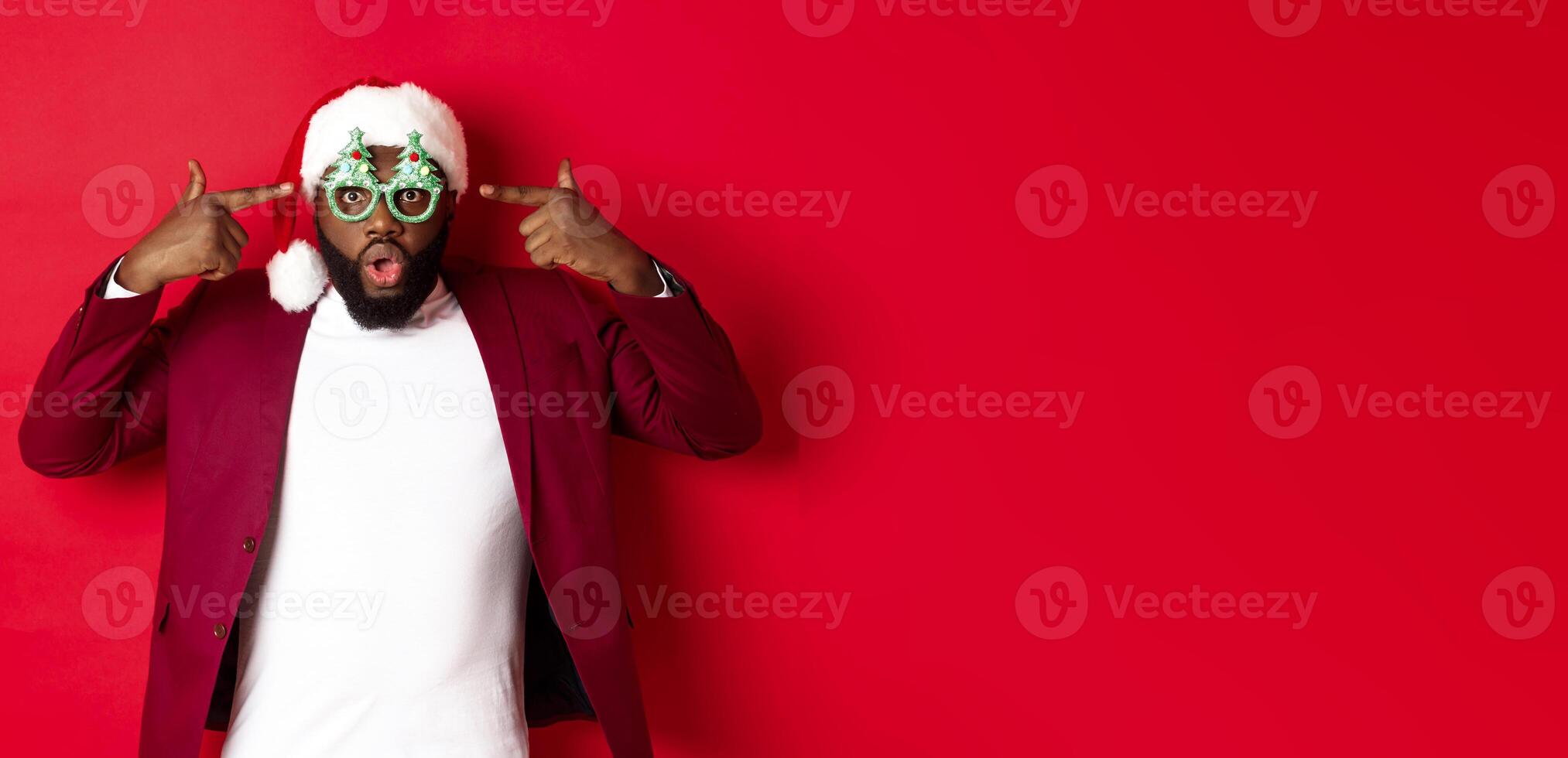Merry Christmas. Cheerful Black man wearing funny party glasses and santa hat, smiling joyful, celebrating winter holidays, standing over red background photo