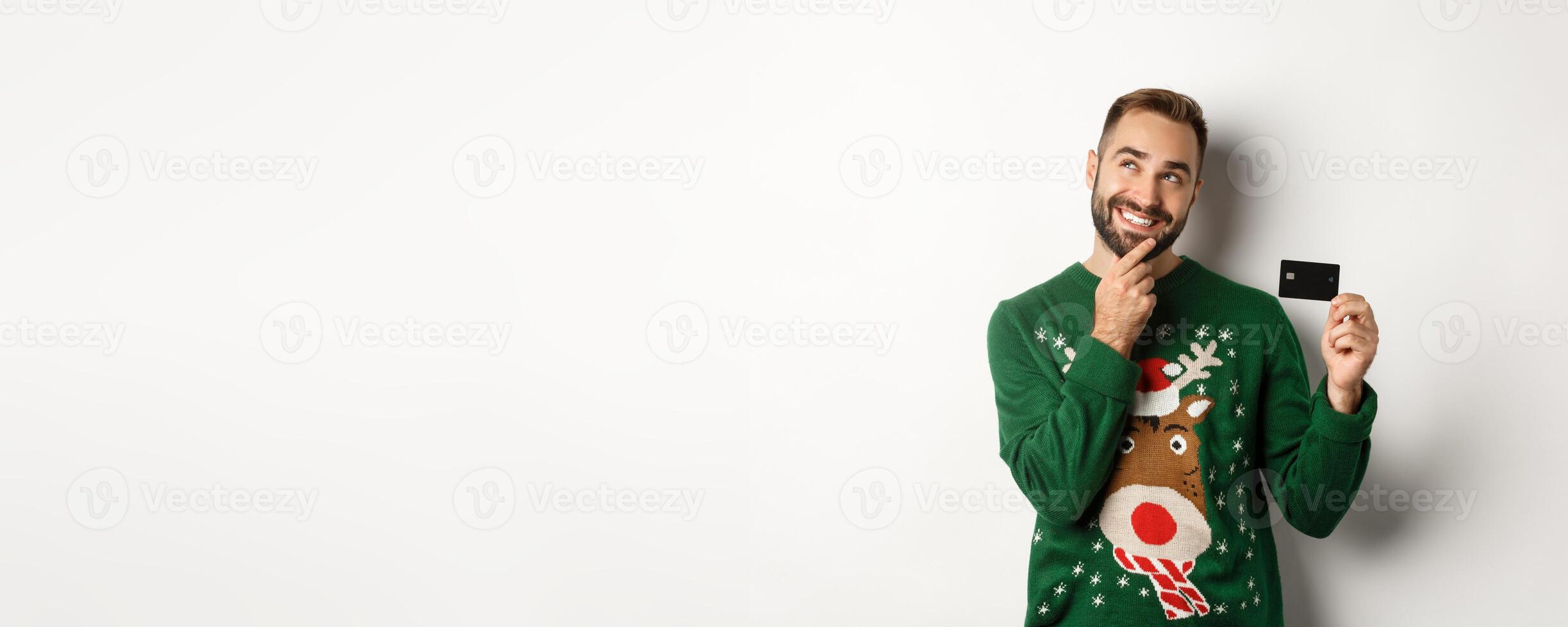 Christmas holidays and shopping concept. Thoughtful man planning New Year gift list, holding credit card and smiling, standing over white background photo