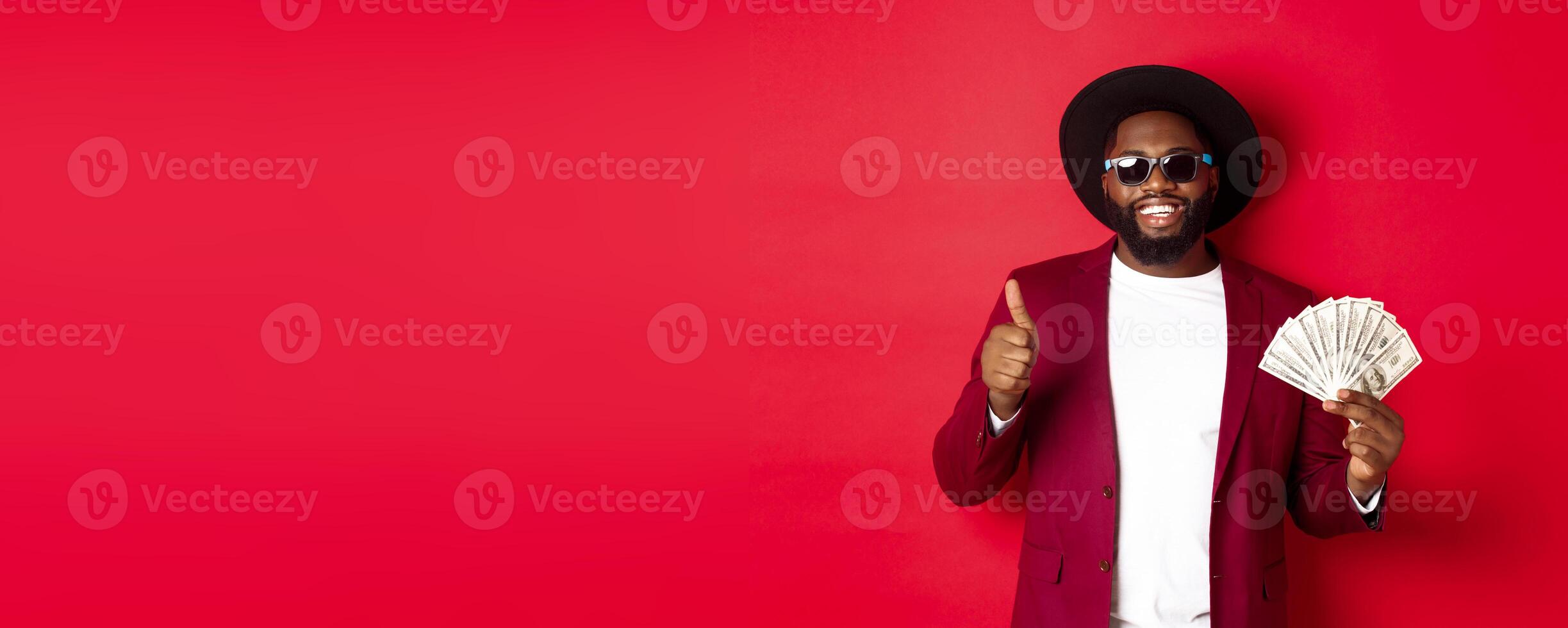 Handsome modern african american man in sunglasses and party clothes, showing thumb up with dollars, earn money and looking satisfied, red background photo
