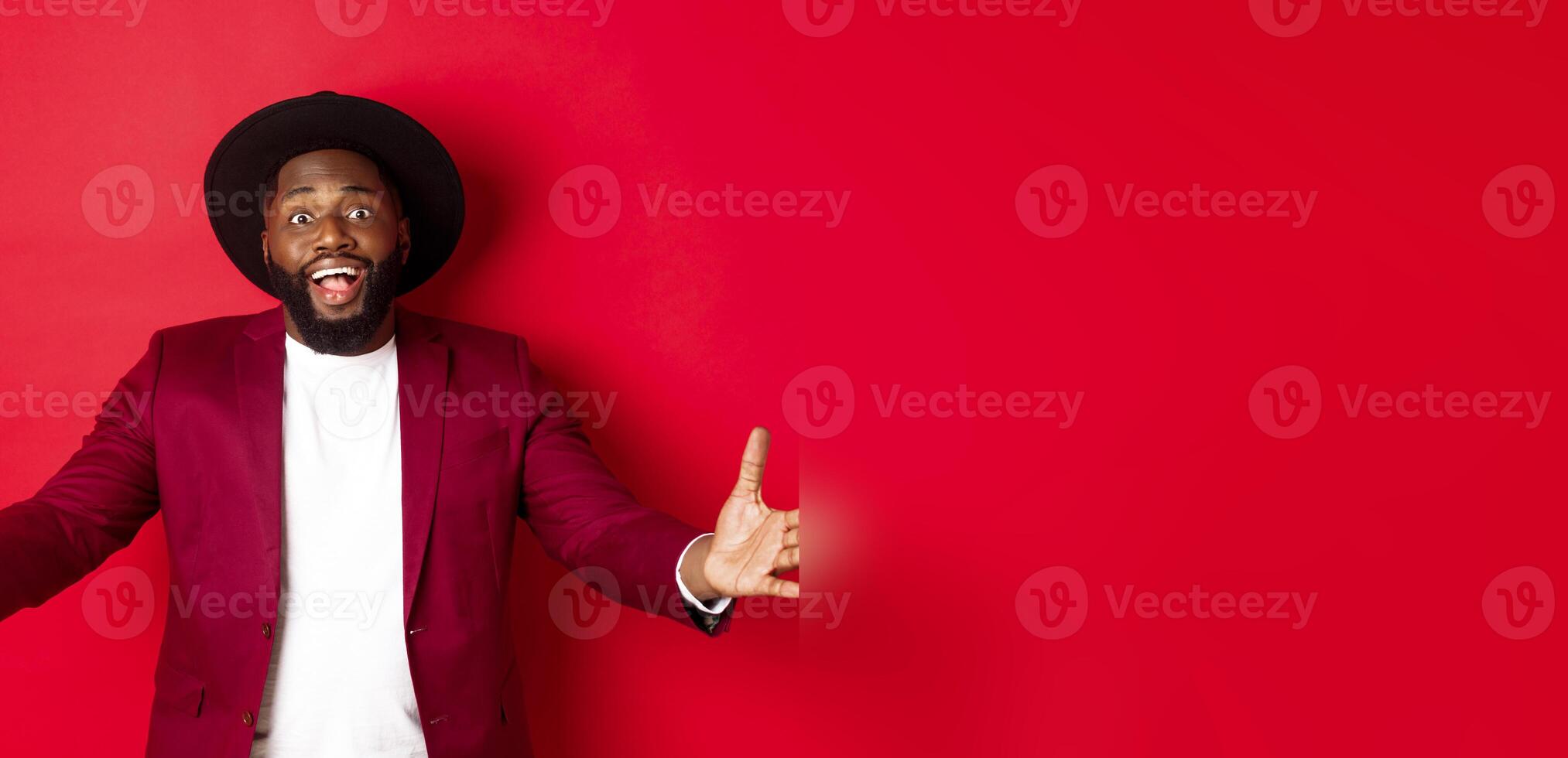 Fashion and party concept. Pleased handsome Black man spread hands in welcome gesture, reaching to receive something, looking amazed, standing over red background photo