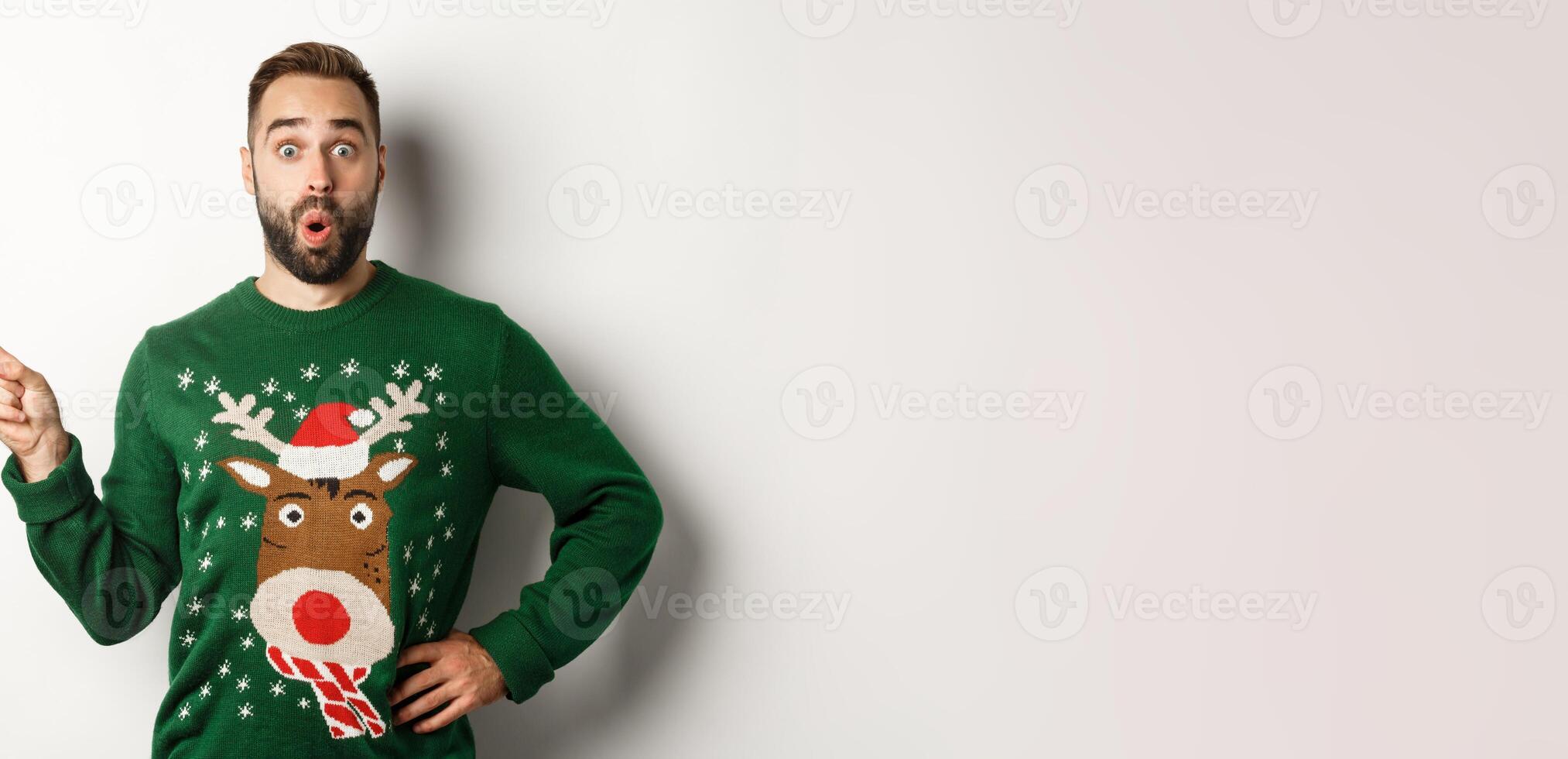 New year celebration and winter holidays concept. Surprised man in christmas sweater pointing finger at upper left corner, looking amazed, white background photo