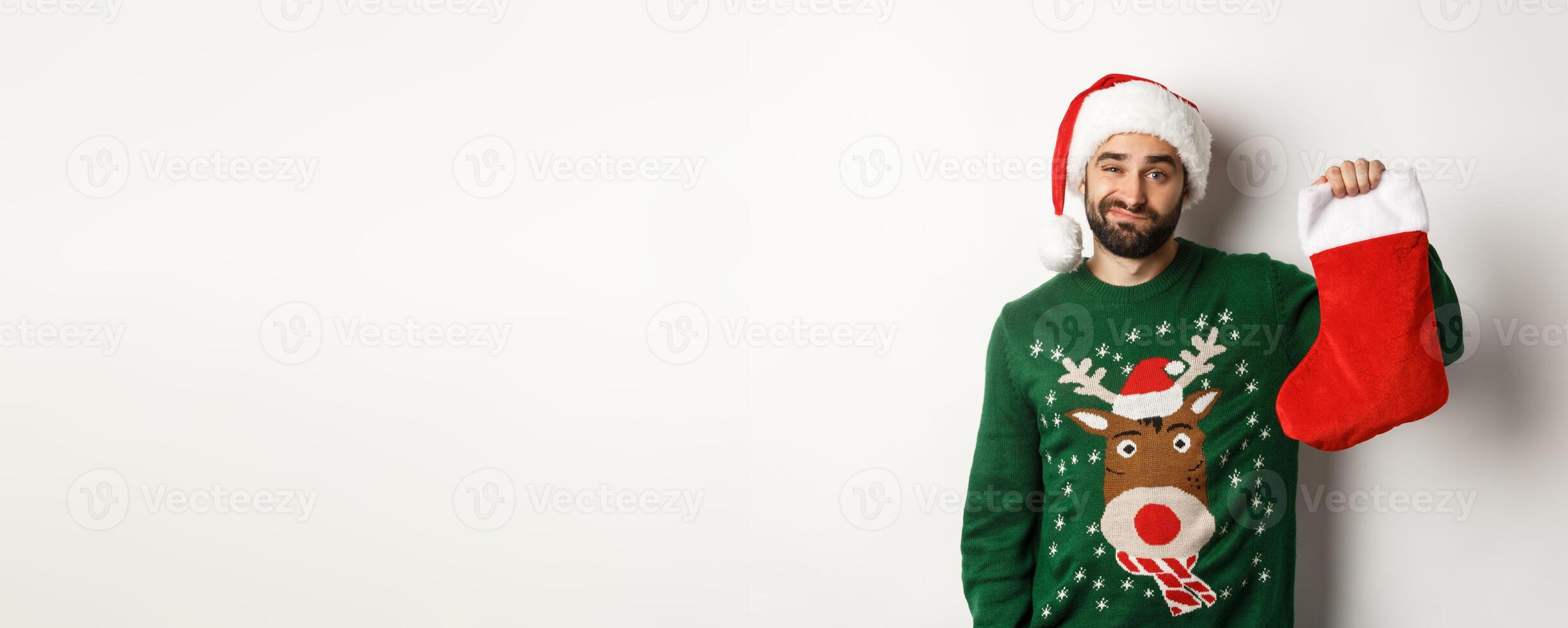 Xmas and winter holidays concept. Displeased guy disappointed in gift in Christmas sock, standing upset in Santa hat against white background photo