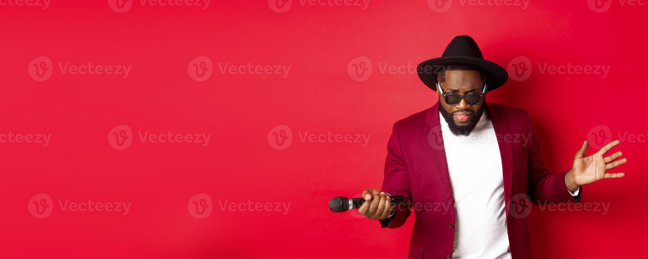 Passionate black male singer performing against red background, singing into microphone, wearing party outfit, standing over red background photo
