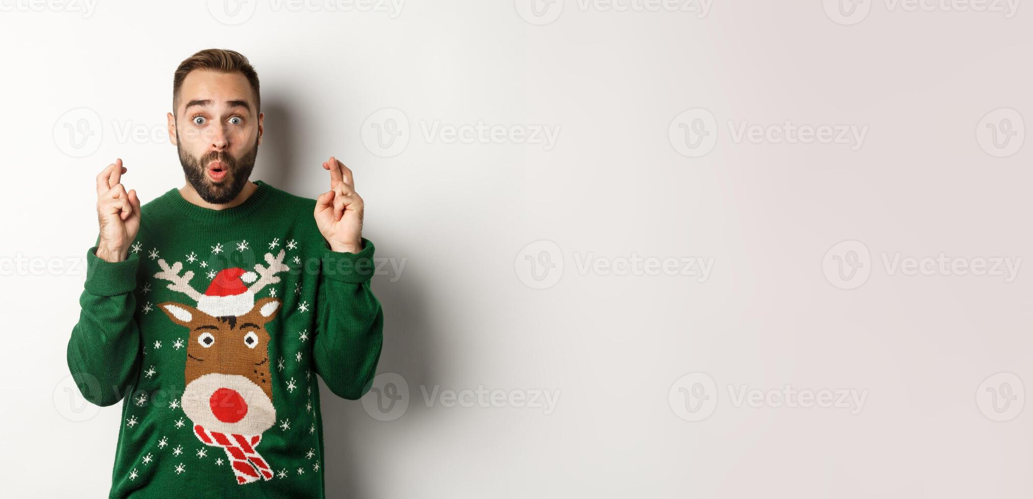 Christmas, holidays and celebration. Excited guy making wish, keep fingers crossed for good luck and smiling, standing over white background photo
