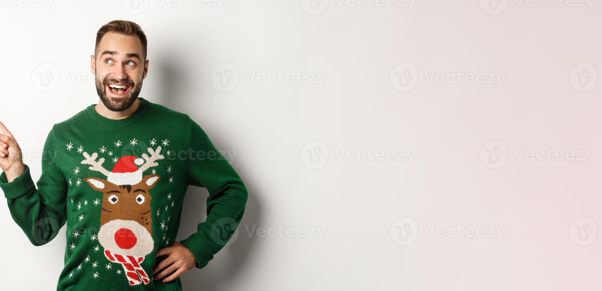 New year celebration and winter holidays concept. Dreamy bearded man in green christmas sweater, pointing at upper left corner and smiling amused, white background photo
