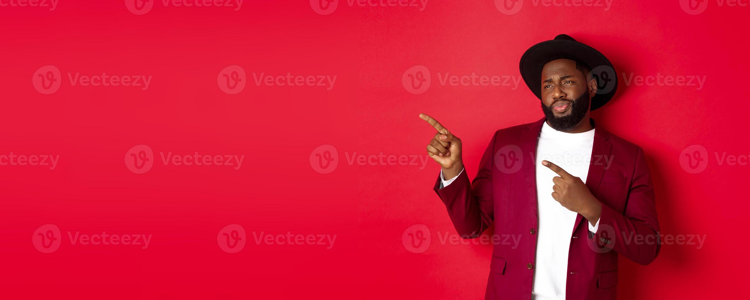 Winter holidays and shopping concept. Skeptical and displeased Black man looking with disdain and pointing finger left at copy space, standing against red background photo