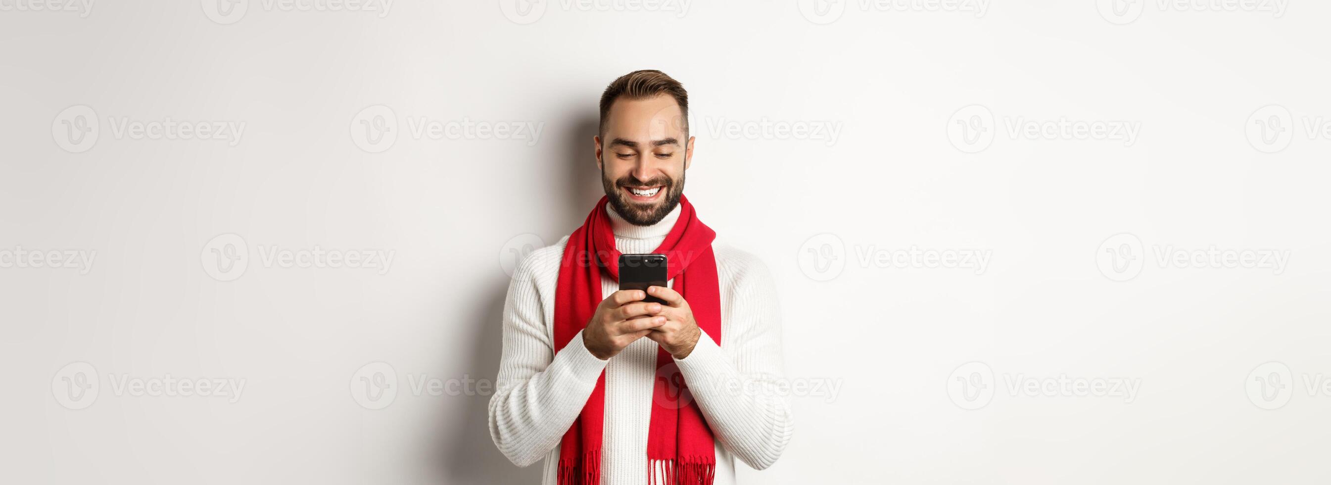 Happy man smiling while reading message on mobile phone, standing in winter sweater and red scarf against white background photo